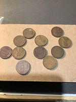 Top view of 12 Lincoln Wheat pennies on a cardboard surface showing obverse and reverse details with legible dates and coin conditions.