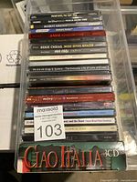 Photo shows 19 music CDs stacked in a plastic container, titles include Miley Cyrus Breakout, Adele 21, Classic Queen, Beauty and the Beast soundtrack, Pocahontas soundtrack, and Ciao Italia 3 CD set. Cases mostly intact with some shelf wear and labels.