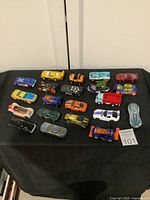 Twenty mixed brand die-cast toy cars arranged on a black cloth background in rows. Includes various colors and types, such as race cars, trucks, and fantasy vehicles.