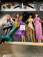 Five fashion dolls lying down, showing a variety of outfits including a mermaid tail and princess dresses, different hair colors and styles.
