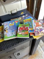 Photo showing 9 children's books featuring Disney, joke, and puzzle titles arranged on a table with a closed plastic toolbox behind.