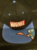Front view of black and blue baseball cap with Marvel logo patch on front panel and Captain America graphic on blue brim.