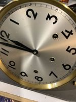 Close-up photo of the metal round clock face showing Arabic numerals, winding holes and black hands with brass rim.