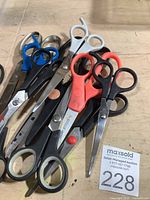 Top-down photo of 9 assorted scissors in a pile showing various handle colors: black, blue, grey, red. Some blades show minor wear and slight staining.