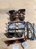 Front view of 8 eyeglasses and sunglasses arranged in rows on a beige surface, showing variety of frame styles and lens colors