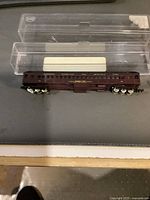 Top-down view of maroon/burgundy model train passenger car with gold lettering and white wheels on a black surface, with original clear plastic case.