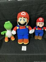 Set of three Nintendo plush toys displayed on a black background, two Mario plush toys of different sizes and one Yoshi plush toy.