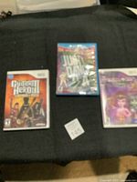 Three Nintendo Wii video games on black fabric, showing Guitar Hero III, Just Dance 2, and Barbie game cases.