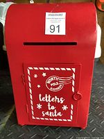 Front view of red metal Letters to Santa mailbox showing the slot and door with white holiday text and lock.
