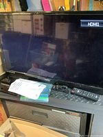 Black 32-inch flat screen TV displaying HDMI1 on screen, placed on a metal surface with remote, manual, and power cord in front.