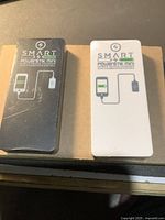 Photo showing both SMART Powerstik Mini portable chargers, one black and one white, on a cardboard surface. Each has device charging graphic and brand markings.