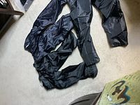 Three black sports pants laid out on a carpet floor showing length and shape. Pants are smooth outer material with elastic cuffs and waistband.