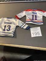 Two miniature hockey jerseys displayed side by side on a grey surface, with auction lot card visible. Blue and white Maple Leafs jersey labeled 'Sundin 13' on the left, red, white and blue Canadiens jersey labeled 'Koivu 11' on the right.
