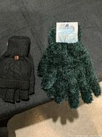 Two pairs of gloves laid on a dark surface. Left pair: black knit fingerless mittens with cable pattern and brown button on cuff. Right pair: dark green fuzzy gloves with soft texture, still attached to product label indicating winter gloves.