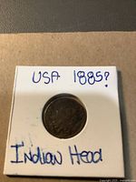 Front side of the USA Indian Head penny in coin holder with handwritten label indicating 'USA 1885? Indian Head'. The coin shows significant wear obscuring details.