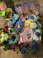 Top view photo showing a variety of vintage Disney Polly Pocket playsets, with colorful miniature buildings, furniture, and pools. Figures including Genie prominently visible. Several small parts appear to be bagged separately within the container.