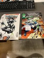 Photo showing two comic books laid flat on a table with a black keyboard partially visible at the top. Left comic is Wildstorm: Phantom Guard #1 with a gray-white cover featuring a crouching female character in armor. Right comic is ShadowHawk #4 with an action-packed cover of a white and blue armored character leaping in a cityscape with green glow effects. Both comics are in plastic sleeves.