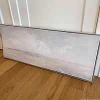 Full view of abstract landscape painting lying against a wall on wooden floor, showcasing horizontal composition with soft neutral tones.