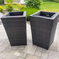 Two tall outdoor planters with dark grey woven rattan-style exterior placed on patio pavers with green shrubbery background.