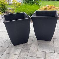 Two square tapered black plastic planters outside on patio pavers with garden plants behind.