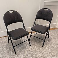 Two black folding chairs shown side by side, both open and standing on carpet. Cushions on seats and backs appear in clean, good condition with ribbed texture fabric covering. Frame is metal with handle on backrest.