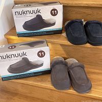 Two boxes of Nuknuuk men's slippers size 11 with two pairs outside the boxes, one dark gray and one navy blue