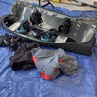 Overview of two snowboards with bindings lying in a soft case on a tarp. Several winter clothing items including jackets and gloves are in front of the case.