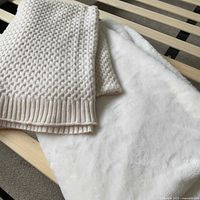Two throw blankets laid out: cream knit textured blanket folded on top of soft white plush blanket on wooden slats.