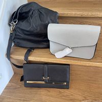 Three purses displayed on wooden stairs: a black leather hobo-style purse, a white structured purse, and a black wallet-style purse.