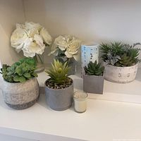 Shelf display of white artificial roses, multiple artificial succulents in decorative pots, and two small white candles.