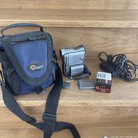 Full view of the lot including Lowepro carrying case, Canon Optura 300 camcorder, battery pack, battery charger with cords, two video cassette tapes, and lens cap.