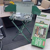 Full view of Coleman propane camp stove in green with wind shields, mounted on matching green foldable stand. Two original packaging boxes beside stand.