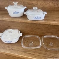 Photo showing three Corningware dishes with blue cornflower pattern, two with glass lids on, and three glass lids placed in front for display.