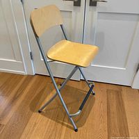 Folding chair with light wood seat and backrest supported by metal tubular frame shown on wooden floor.
