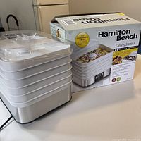 Front view of the Hamilton Beach food dehydrator next to its original box, showing 6 white trays stacked and clear lid.