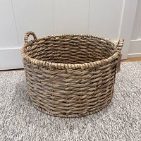 Full view of large round woven basket with braided rim and loop handle on gray carpet.