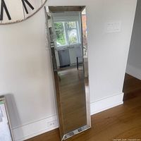 Full view of tall rectangular wall mirror leaning against the wall in a room, showing the beveled mirror frame reflecting the surroundings.