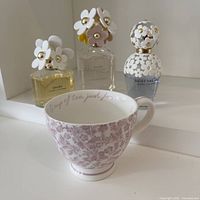 Full view showing three Marc Jacobs Daisy perfume bottles in front of a mirror and a pink floral ceramic tea cup.