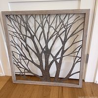 Full front view of square metal tree wall art with wooden frame leaning against a wall and door.