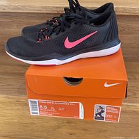 Photo of black Nike Flex Supreme TR5 running shoes placed on top of the original orange Nike shoe box. Shows shoe profile, Nike swoosh in pink, shoe laces, and sole.