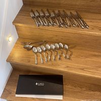 Photo showing all cutlery pieces arranged on wooden stairs: 12 forks, 6 spoons, 1 serving spoon, 1 serving fork, 1 sugar tongs, and Cutco box at the bottom