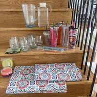Overall view of glassware including acrylic tumblers with lids and straws, clear pitcher, stemless wine glasses, packaged items, and folded patterned tablecloth with placemats on wooden stairs.