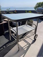 Full view of the patio caddy showing rectangular textured glass top, tubular metal frame, two wheels on one side, and legs on the other.