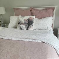 Bed with queen quilt in light pink with floral texture and ruffled edges, white fitted sheet with small floral pattern, and multiple pillows including pink striped shams, floral decorative pillow, and white pillows with subtle pattern.