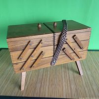 Front view of the closed vintage wooden sewing box on legs with folding parts and bird's eye knobs.