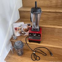 Overall shot of red Vitamix base with two pitchers, tamper, recipe books and accessories on wooden stairs