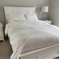 Photo of a made bed showing the white double duvet cover and four matching pillows in a textured, quilted pattern on a white wooden bed frame.