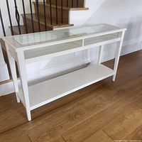 Full view of the white side table showing the glass inset top, open compartments underneath, and the lower shelf against a wooden floor near stairs.
