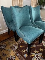 Four teal upholstered dining chairs grouped together on a patterned rug, showing front and partial side views.