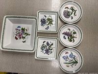 5 botanical plates arranged on a table: 1 larger square, 2 smaller squares, and 2 round plates, each featuring botanical flower and butterfly designs with green leafy borders.